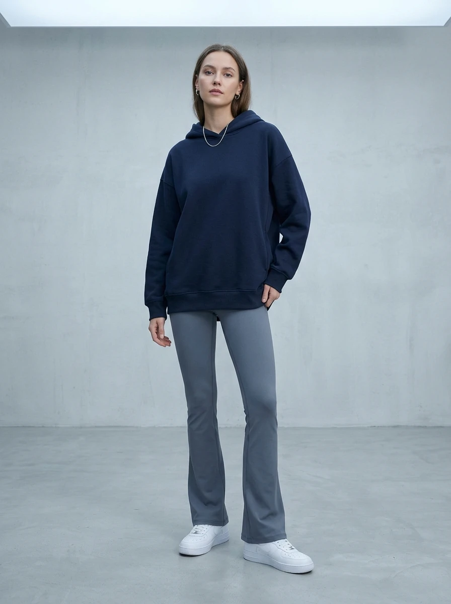 A woman stands in a studio wearing slate gray flare leggings and a navy blue hoodie with white sneakers.